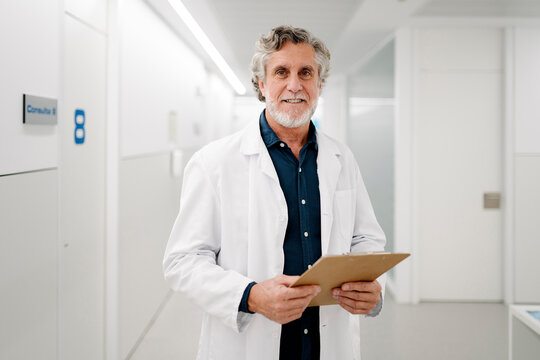 Senior doctor holding clipboard smiling in hospital corridor