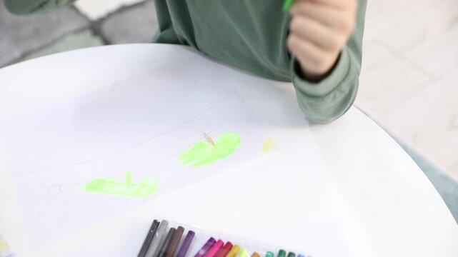 Little boy drawing with green felt pen at white table indoors, above view
