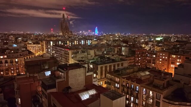 Night aerial view of Barcelona showing the illuminated Sagrada Fam&iacute;lia rising above dense Eixample rooftops, with Torre Gl&ograve;ries glowing blue on the horizon, slow push in aerial.