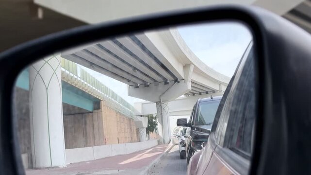 Car side mirror view shows slow traffic queue beneath an elevated highway overpass, with multiple vehicles lined up on an urban road in bright daytime light.