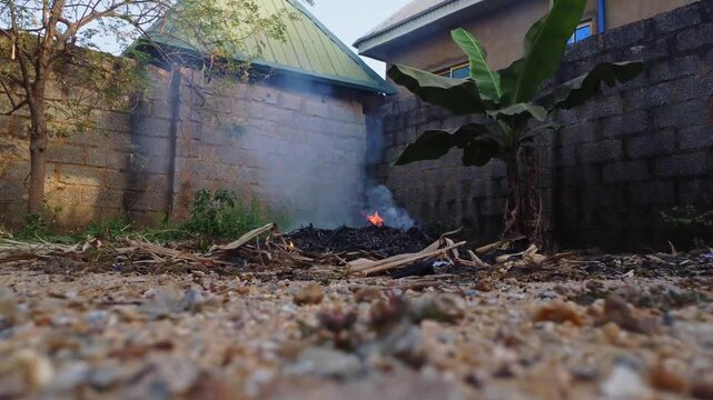 Low angle view of a burn pit with flames and a lot of smoke in a garden in Nigeria, Africa. Banana and moringa tree are seen behind