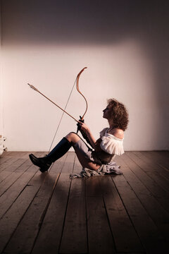 Person aims bow while sitting on wooden floor in a room