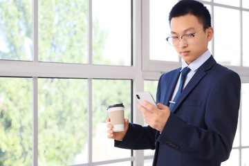 Fototapeta premium Chinese businessman with coffee using mobile phone near window in office