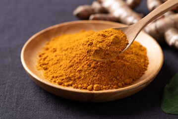 Turmeric powder in a wooden bowl with a wooden spoon scooping it, and fresh turmeric roots on a...