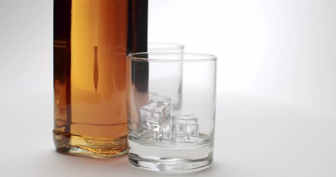 Golden whiskey bottle with two elegant glasses filled with ice cubes slowly rotating on a clean, isolated white background, showcasing the amber color of the spirit and the classic bar setup.