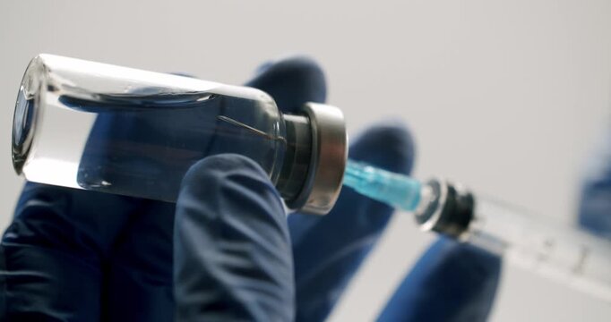 Medical professional in blue sterile gloves drawing a clear liquid vaccine from a glass vial into a syringe, preparing for an injection on an isolated white background in a hospital