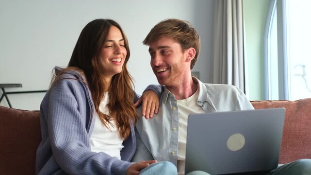 Happy millennial couple sit relax on couch in living room watching video on laptop together, smiling young husband and wife rest on sofa at home browsing Internet using modern computer device

