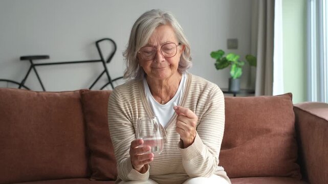 Close up senior mature woman taking painkiller, antibiotic, supplements or vitamin medication, middle aged female holding glass of water and white round pill in hands, disease treatment concept
