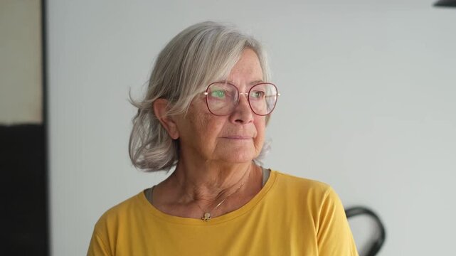 Elderly woman looks out the window with a worried expression. She reflects on her thoughts and feels concerned about her day while standing at home.
