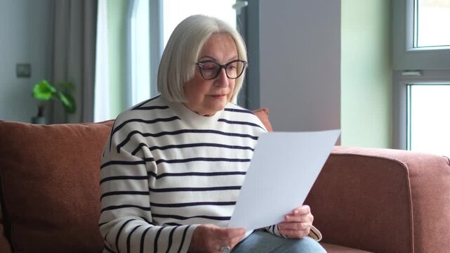 Focused elderly 60s woman sit on sofa in living room reading postal correspondence or bank notice, concentrated mature 50s female rest on couch at home consider message in paper post letter
