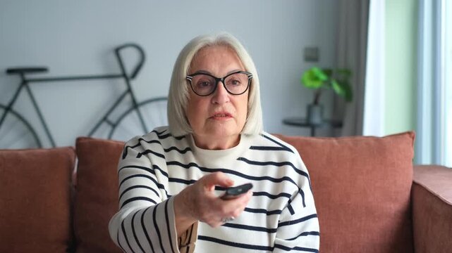 Happy smiling senior woman sitting on couch in the living room, pointing tv remote controller to switch channels, watching favorite movie online series
