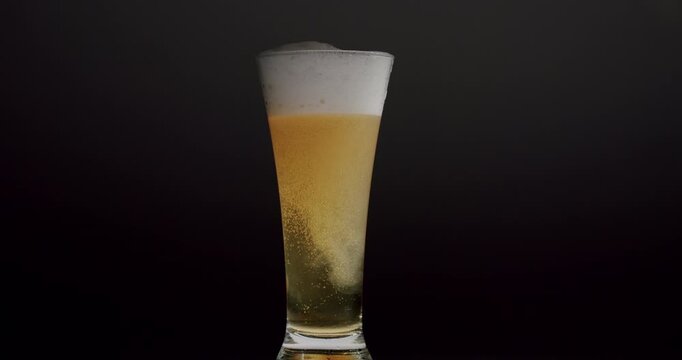 Cold light beer in a glass with bubbles and swirling sediment creating patterns against a dark studio background. The frothy drink is a refreshing alcoholic beverage perfect for a party or bar