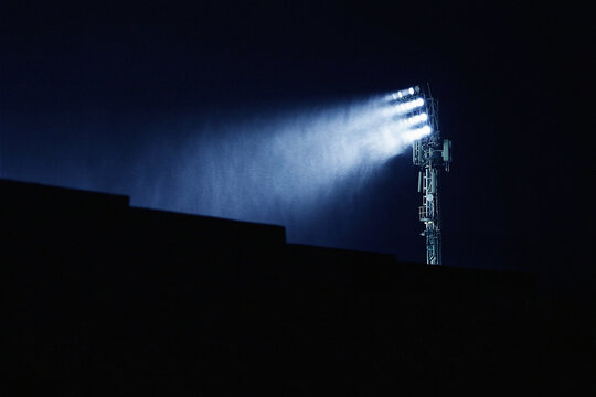 A tall stadium floodlight tower illuminates a dark sports arena at night, with powerful beams of light cutting through mist and rain. The silhouette of a stadium stand creates a dramatic foreground ag