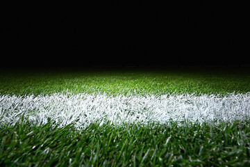 A close-up view of a vibrant green football pitch with a crisp white line, illuminated by stadium lights at night. This low-angle shot captures the detailed texture of the grass and the precision of t