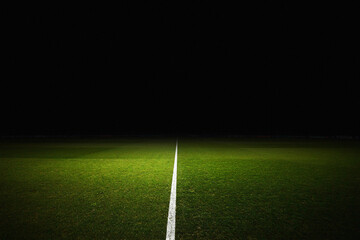 Professional soccer field at night with a bright spotlight illuminating the green grass and white center line. The dark background features blurred advertising boards for major sponsors, creating a dr © Pix
