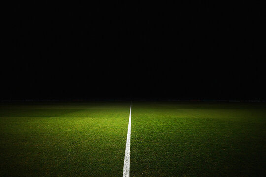 Professional soccer field at night with a bright spotlight illuminating the green grass and white center line. The dark background features blurred advertising boards for major sponsors, creating a dr