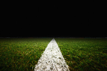 A low-angle, close-up shot of a white boundary line on a professional soccer pitch at night. The detailed green grass texture leads the eye toward a dark background where blurred stadium lights and ad © Pix