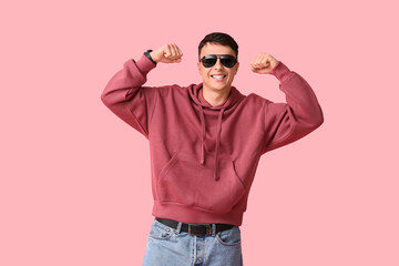 Fototapeta premium Handsome young man in stylish hoodie and sunglasses showing muscles on pink background