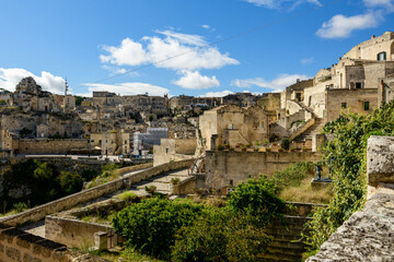 Obraz premium Expansive view of historic stone buildings and terraced rooftops in Matera, Italy, under a clear blue sky with scattered clouds. Sunlight highlights textured limestone architecture and lush greenery