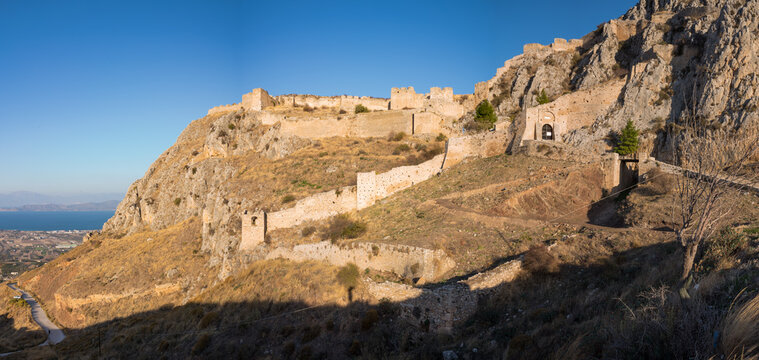 Expansive panorama of Acrocorinth fortress with ancient stone walls winding along a rugged, sunlit hillside, overlooking the plains and distant blue water under a clear sky.