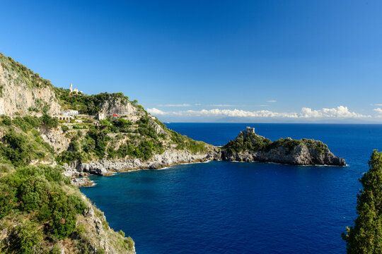 Rocky cliffs covered with lush Mediterranean vegetation rise above the deep blue waters at Capo di Conca, Italy. A historic watchtower stands on a small promontory, framed by vivid skies and sparkling