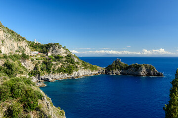 Naklejka premium Sunlit rocky cliffs and lush Mediterranean vegetation frame the historic watchtower on the Capo di Conca promontory, Italy. Deep blue waters of the Amalfi Coast stretch beneath a vivid sky
