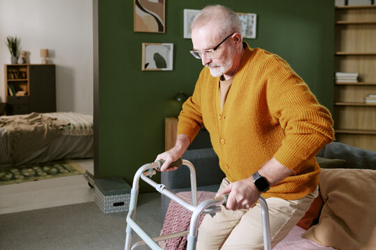 Senior Caucasian man using walker standing up from bed in modern home interior, wearing glasses and smartwatch, demonstrating mobility assistance for elderly independence