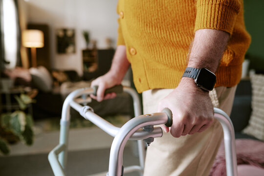 Senior Caucasian man using walker indoors, hands gripping handles while moving forward, wearing smartwatch, partial body visible, home environment in background