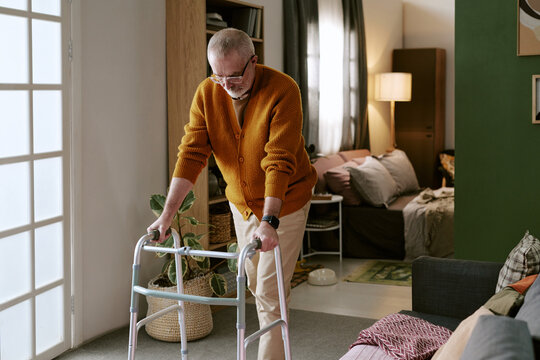 Senior Caucasian man using walker for mobility support in living room, standing and looking down while concentrating on movement, wearing glasses and casual clothing, home environment visible