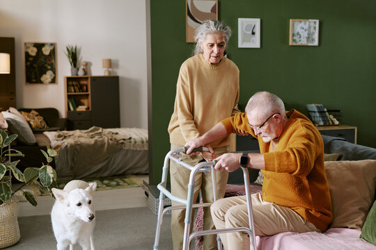 Senior Caucasian woman assisting senior Caucasian man using walker in living room, elderly couple engaging in daily activity with dog standing nearby observing interaction