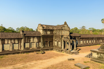Fototapeta premium Historic sandstone gallery with an ornate entrance pavilion lines the second-level courtyard of Angkor Vat, Cambodia. Weathered stonework and geometric rooflines stand out in the warm afternoon sun