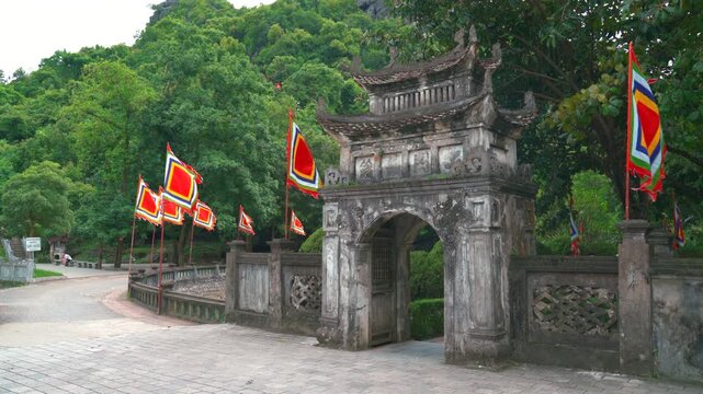 A historic stone entrance gate adorned with vibrant traditional Vietnamese festival flags, leading to the ancient capital complex of Hoa Lu in Ninh Binh.