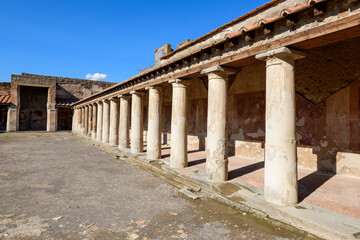 Fototapeta premium Ancient Roman columns line a sunlit portico in the Stabian Baths of Pompeii, Italy. The weathered stone and faded frescoes are highlighted by crisp blue sky, evoking the preserved grandeur of this