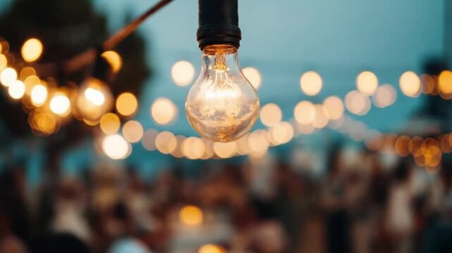 A close-up of a glowing incandescent light bulb from a string of decorative outdoor lights, casting a warm, inviting ambiance. The background features shimmering golden bokeh lights and blurred silhou