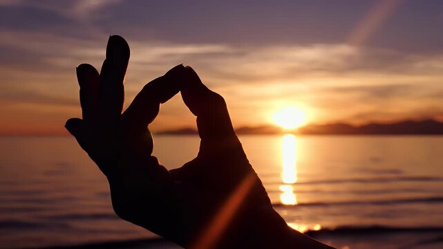 Silhouette of a hand forming a mudra against a vibrant sunset over the ocean
