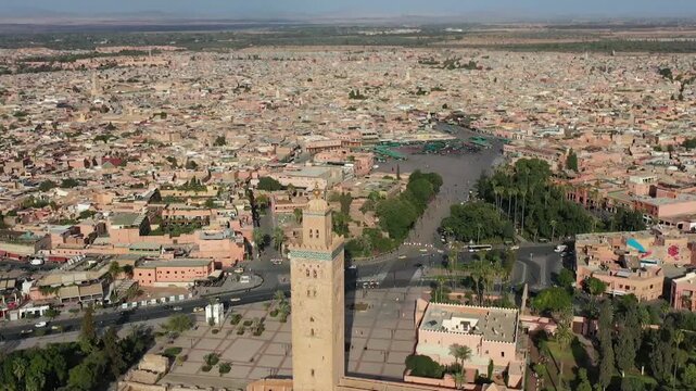 Majestic drone shot featuring the historic Koutoubia minaret. Captures the sprawling red-toned Medina, the distant Jemaa el-Fnaa square, and the traditional architecture of central Marrakech.