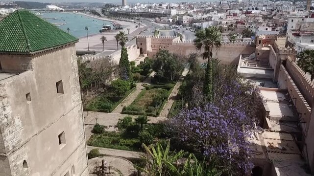 Beautiful drone shot overlooking the Andalusian Gardens within the historic Kasbah of the Udayas. Features the fortified walls, green-tiled towers, blooming jacaranda trees, and river backdrop.