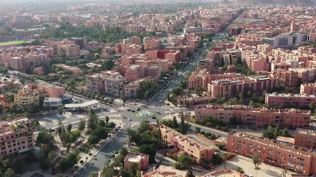 Panoramic drone shot capturing a modern district in Marrakech. Highlights the organized traffic circle, palm-lined avenues, and the iconic red architecture stretching towards the mountains.