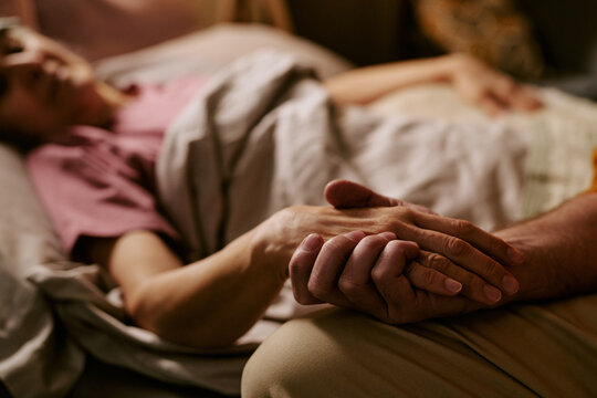 Senior Caucasian woman lying in bed holding hand of senior Caucasian man, showing support and comfort during illness or recovery, focus on hands and connection