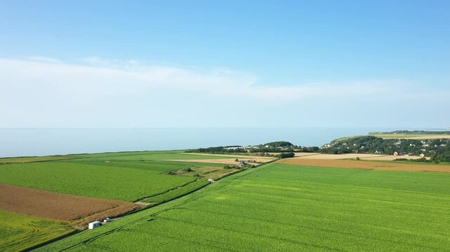 Aerial view of lush green fields and patchwork farmland along the Normandy coast under a clear blue sky. Rural landscape with distant sea horizon and scattered houses.