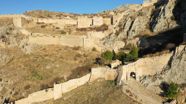 Wide view of the historic stone walls and ruins of Acrocorinth fortress on a rocky hillside in Greece under clear blue sky. The scene captures the rugged terrain and ancient architecture.