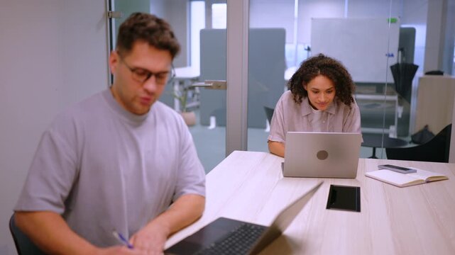 Male employee writes notes beside laptop while female colleague works on computer in background. Bright glass office setting reflects focus, productivity and modern corporate routine.
