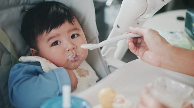 4K slow motion footage of an Asian mother feeding her six month old baby boy with a spoon. Tender parenting moment showing care, bonding, and infant nutrition in a warm home environment.