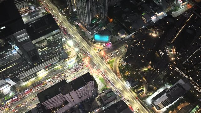 Aerial timelapse view of a busy city intersection at night, captured from a top-down perspective. Streams of car lights move through illuminated roads surrounded by modern skyscrapers, reflecting fast
