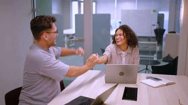 Young coworkers smiling and giving high five across desk with laptops open. Modern office setting reflects teamwork, achievement, startup energy and positive corporate culture.