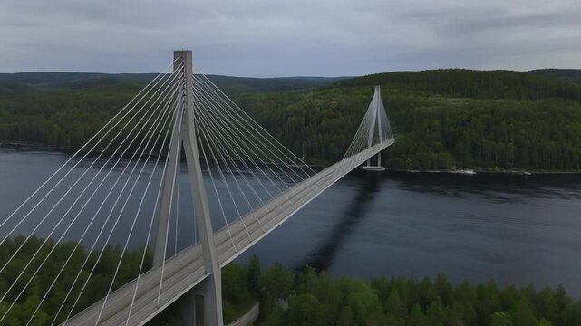 A large cable-stayed bridge spans a body of water, surrounded by dense green forests and under a cloudy sky