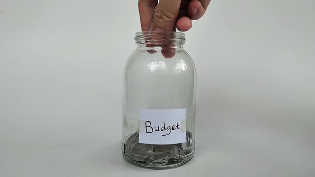 Close-up shot of a hand putting coins or bills into a transparent jar labeled "budget", symbolizing financial planning.