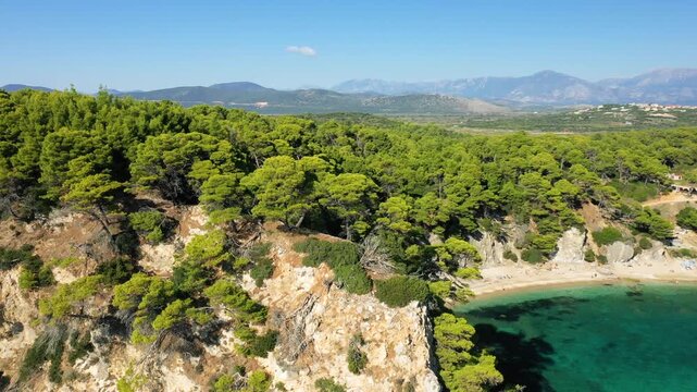 Aerial view of rugged cliffs and dense pine forest overlooking the turquoise waters of Alonaki Fanariou beach in Greece, with distant mountains under a clear blue sky.