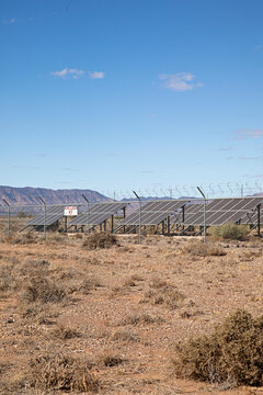 Solar farm in arid, outback Australian landscape with barbed wire fence and distant mountains under a blue sky