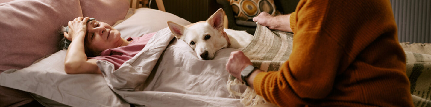 Senior Caucasian woman lying in bed with hand on forehead, dog resting beside her, Caucasian man covering her with blanket, showing caregiving at home